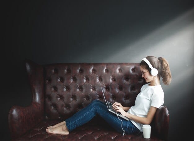 A person sits comfortably on a couch, studying on a laptop with headphones on. Sunlight streams through the window, creating a warm and inviting atmosphere, illustrating the flexibility of online learning.
