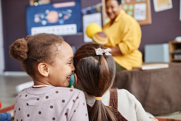 Two young children hugging each other in a classroom setting, showing affection and friendship. Other children are playing and interacting in the background, with a teacher supervising.