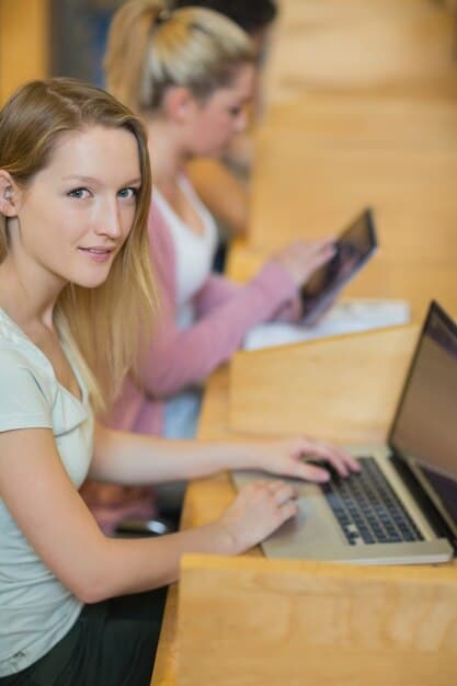 A young student typing on a laptop in a well-lit classroom. There are other students around, and the teacher is in the background, helping another student. The focus is on the student's engagement and the use of technology in education